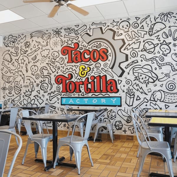A vibrant mural with the Tacos & Tortilla Factory logo and playful Mexican-themed doodles covers a brick wall.