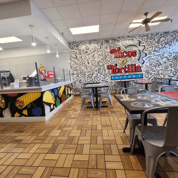 The Tacos and Tortilla Factory dining area features a vibrant mural, light wood-patterned floor, and gray metal tables.