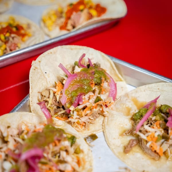 Close-up of three Tacos and Tortilla Factory tacos with meat, slaw, pickled onions, and green salsa.