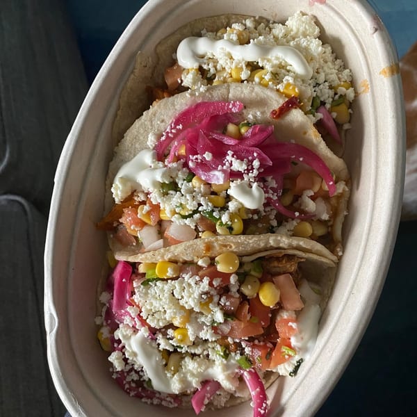 Three Tacos and Tortilla Factory tacos in a container, topped with corn, cheese, and pickled onions.